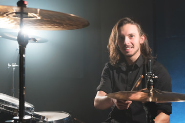 Fototapeta na wymiar Portrait of a smiling long-haired drummer with chopsticks in his hands sitting behind a drum set. Low key. Concepts of the creative freedom of the millenial generation