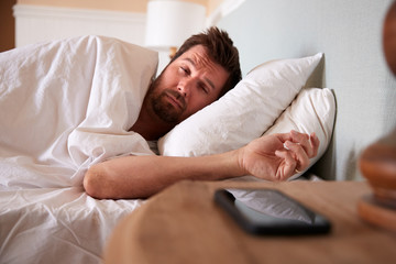 Fototapeta premium Mid adult man asleep in bed, looking at the smartphone on the bedside table in the foreground
