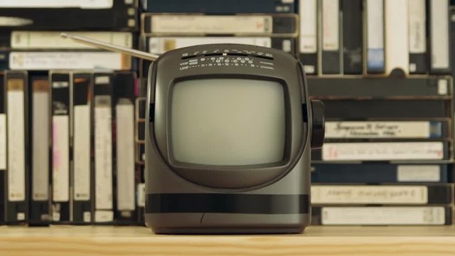 Old small TV set with antenna against background made of VHS and S-VHS video tapes. Camera moving away from TV set and out of focus. Dolly shot