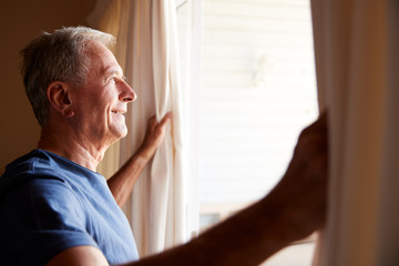 Fototapeta premium Smiling senior white man opening the curtains on a sunny morning, side view, close up