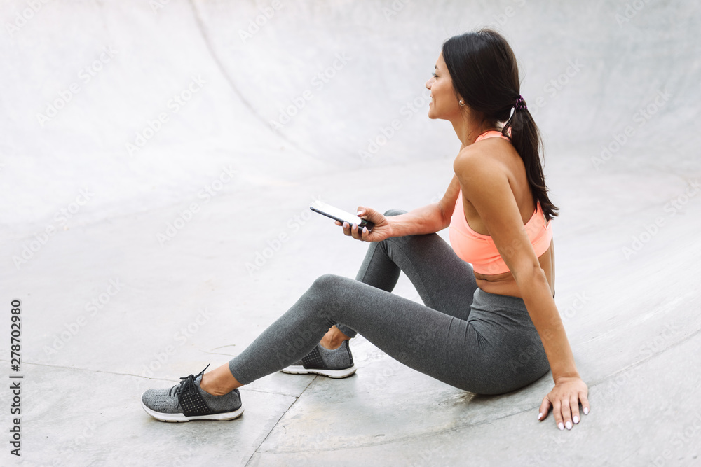 Wall mural Image of happy young woman holding cellphone while sitting on concrete floor outdoors