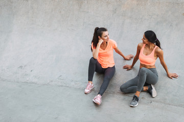 Image of attractive fitness women in sportswear talking together while sitting on concrete sports ground