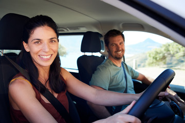 Mid adult white woman driving car, her husband in front passenger seat, smiling to camera, side view