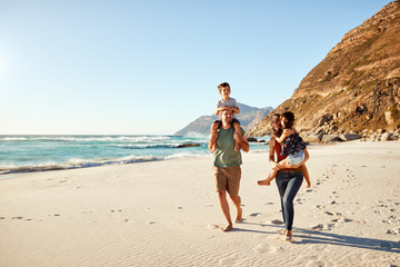 Mid adult white couple walking along on a beach on a family holiday carrying their kids, full length - Powered by Adobe