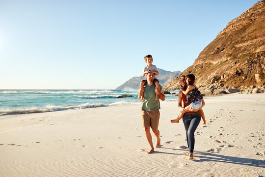 Mid adult white couple walking along on a beach on a family holiday carrying their kids, full length