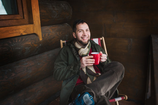 Happy man drinks tea sitting in chaise lounge in the winter wooden chalet dressed in knitted sweater
