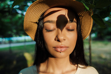 Portrait of relaxed woman wearing straw hat poising with eyes closed and shadow of leaves at her face in green park