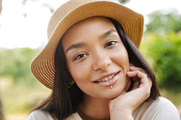Photo of joyful woman wearing straw hat smiling at camera while walking in green park