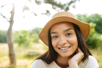 Photo of cheerful woman wearing straw hat smiling at camera while walking in green park