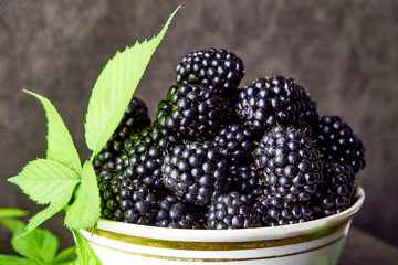 Ripe blackberries with leaves in a bowl on old wooden boards on a dark background.