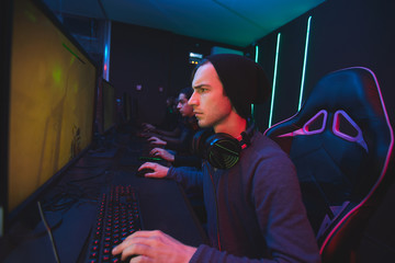 Fototapeta premium Serious young man in beanie sitting at table and using computer while playing shooter video game