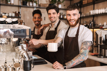 Three cheerful male baristas standing at the coffee shop - obrazy, fototapety, plakaty