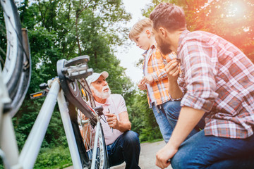 Senior man helping his son and grandson fixing upside down bike outdoor in city park in summer day. Family relation different generation concept.