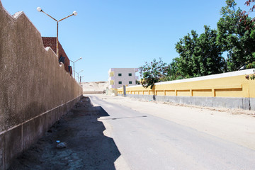 Fototapeta na wymiar Street with no people in sunny day in suburban Hurghada, Egypt. There are beach, wall with rough surface, yellow wall, woods, brick house, and old apartment in image.