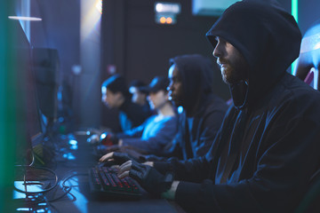 Group of serious gloomy young hackers in hoodies sitting in row and typing on keyboards fastly in...