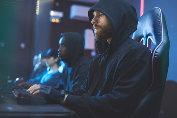 Serious geek guy with beard wearing black hoodie sitting in comfortable chair and searching for information on internet while using computer