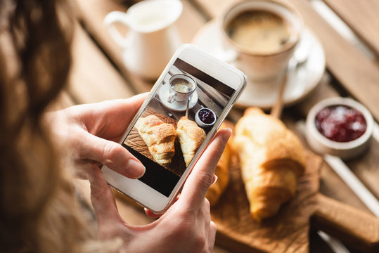 Woman taking picture of breakfast in cafe. Croissant, coffee with cream and jam on wooden table. Toned image, lifestyle and social media concept