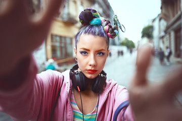 Playful cool funky hipster young girl with headphones and crazy hair taking selfie on street - 278718193