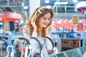 Red-haired girl standing in front of the stand in the electronics store chooses headphones before...