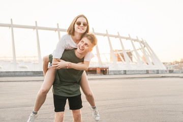 Young happy lovers couple in the urban landscape, young man holds the girl on her back. Girl hugs the boy on the street. Stylish couple having fun on the urban background. Lifestyle