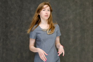 Portrait to the waist of a young pretty brunette girl woman with beautiful long hair on a gray background in a gray dress. He talks, smiles, shows his hands with emotions in various poses.