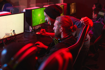 Positive excited hipster guys playing computer game together in modern pc game club