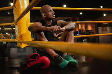 Fototapeta premium Attractive young male African male boxing fighter resting after fighting in boxing rink, copy space. Handsome strong boxer concentrating before his fight