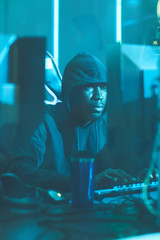 Serious young Afro-American hacker in hoodie sitting at table and typing on computer keyboard while composing data virus code