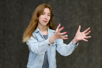 Waist up portrait of a young pretty brunette girl woman with beautiful long hair on a gray background in a jeans jacket. He talks, smiles, shows his hands with emotions in various poses.