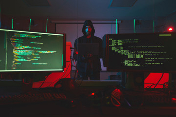 Serious young bearded hacker in hoodie standing in server room with computers and using laptop while getting into computer system