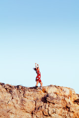 Fototapeta premium Young girl with red dress on cliff's edge with raised hands and looking to a sky