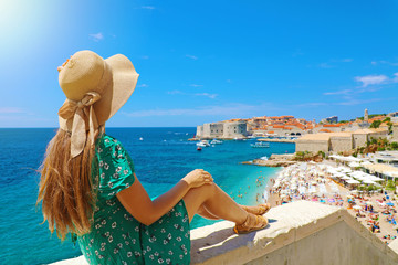 Beautiful young woman with hat sitting on wall looking at stunning panoramic village of Dubrovnik in Croatia, Europe