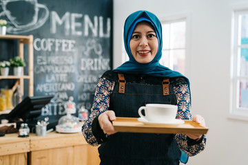 young muslim employee barista woman standing holding cup of tea on plate at coffee shop counter....