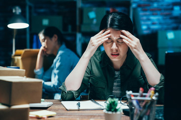 Stressed young businesswoman working late in dark office at night frowning with hands cover massage forehead hurting head headache. colleague sleeping in e commerce online shop full with parcel boxes
