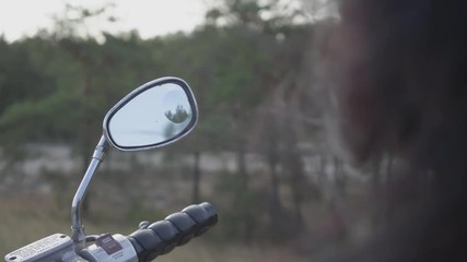 Back view of unrecognizable girl sitting on the motorcycle cleaning the mirror with her gloved hand. Hair fluttering in the wind. Hobby, traveling and active lifestyle. Female biker outdoors