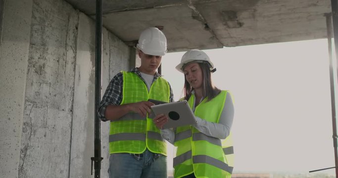 Engineers designers stand on the roof of the building under construction and discuss the plan and the progress of construction using a tablet and mobile phone