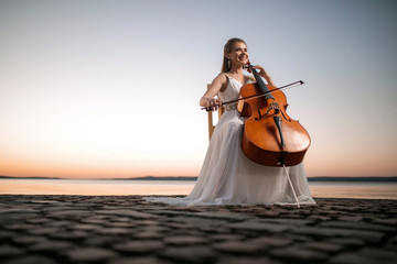Girl in a white dress playing the cello on the shore of the lake, after sunset. There is a place...