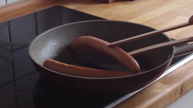 A person turning frankfurter sausages in a home frying pan while cooking lunch in the kitchen.
