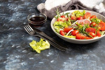 Salad of tomato, onion, pepper, arugula and chia seeds. Dressed with soy sauce and olive oil. Near the stick with dressing. On a dark background. Metal fork and knife. 