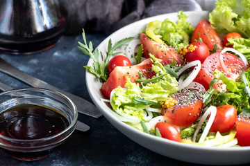 Salad of tomato, onion, pepper, arugula and chia seeds. Dressed with soy sauce and olive oil. Near the stick with dressing. On a dark background. Metal fork and knife. 