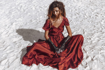 beautiful young fashionable woman in elegant dress sitting on sand at sunset