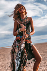 young boho style woman sitting on sand on the beach at sunset