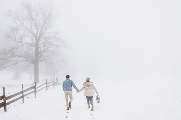 Rear of the young Caucasian couple in love holding hands and running during a blizzard in the countryside and girl holding a lantern. Back view.