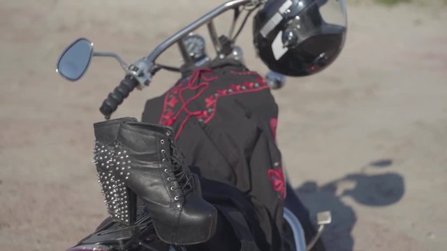 Spiked shoes with high heels and black and red dress lying on the motorcycle close-up. The motorbike with the helmet on the wheel on the riverbank. Hobby, traveling and active lifestyle