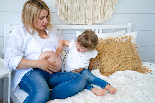 Pregnant Woman With Her Little Daughter On The Bed In Her Bedroom. The Girl Listens To The Baby In Her Mother's Stomach. Waiting For Addition In The Family