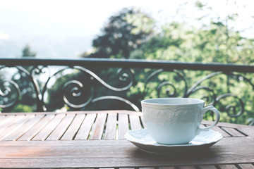 White ceramic cup of tea on a saucer on wooden table against picturesque background. 