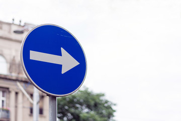 Right direction allowed one way blue street sign on a city street, against the gray buildings and cloudy sky bokeh background