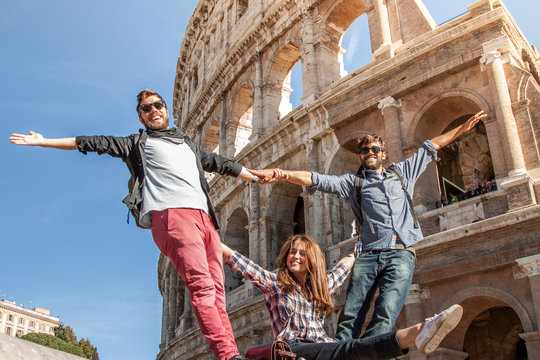 Three happy young friends tourists standing in front of colosseum in rome posing for pictures having fun