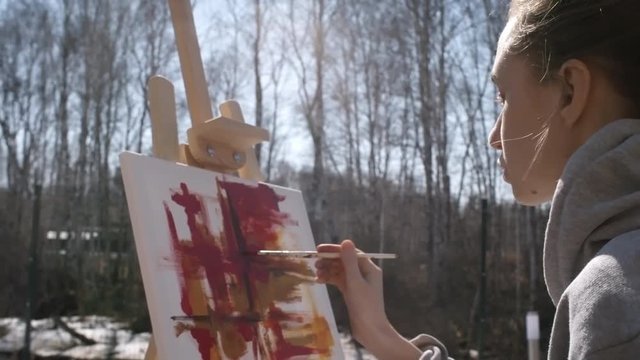 Handheld tilt down shot of female painter sitting in the woods with snow on ground, holding palette and drawing abstract painting with oil paint on canvas