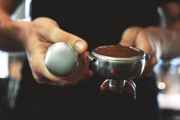 barista man holding coffee holder with ground coffee in one hand and a tamper in another standing near professional coffee machine preparing americano in cafe close up